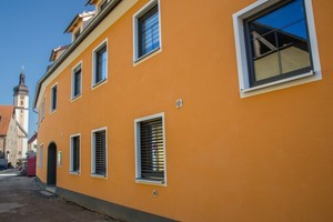  Das neue Gebäude (links) ist wie der Altbau (rechts) städtebaulich in einer klaren Linie der Straße entlang gezogen worden. Im Hintergrund ist die Pfarrkirche Maria Himmelfahrt mit ihrem schlanken Turm zu sehen 